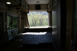 Step inside this RV rental: a comfy bed, kitchen area, and a large rear window offering stunning views of green trees and mountains.