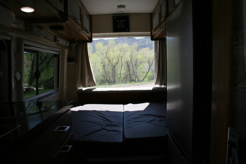 Step inside this RV rental: a comfy bed, kitchen area, and a large rear window offering stunning views of green trees and mountains. — photo 11
