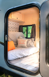 Cozy RV rental camper interior: light wood, bed, window, and a unique orange accent pillow.