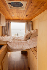 Inside a cozy wooden rv rental, a comfortable bed offers direct views of the ocean & a surfer on the beach. Van life comfort.