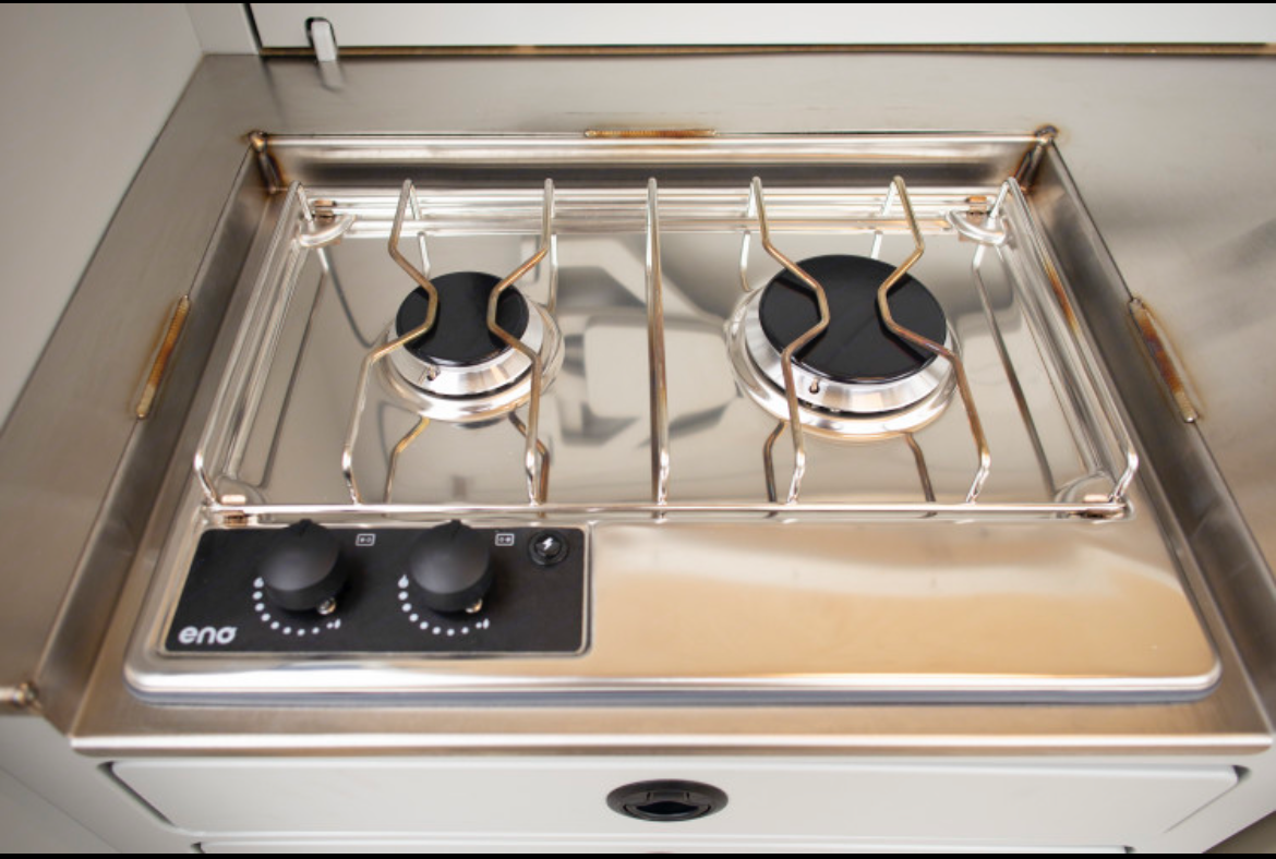 Inside an RV rental, a compact stainless steel two-burner gas stove features black control knobs for cooking ease.