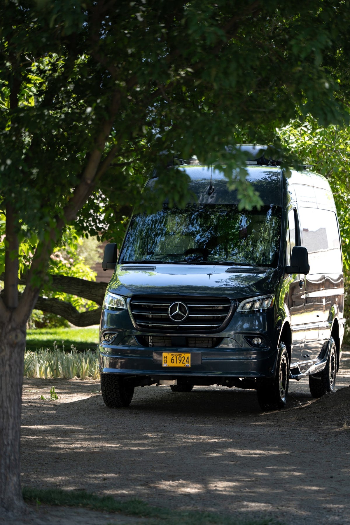 Dark grey Mercedes Sprinter van rv rental with Oregon plate 61924, parked on a shaded dirt path under lush green trees. — photo 5