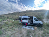 Adventure-ready RV rental: Silver camper van with open doors, dog, and camping setup on a scenic, brush-covered hill under cloudy sky.