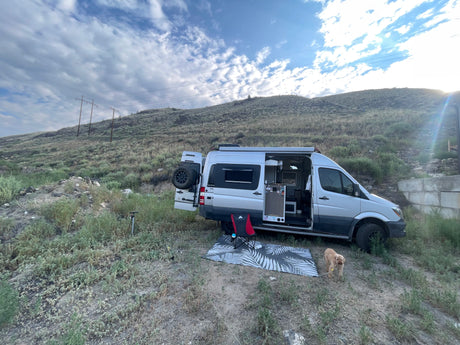 Adventure-ready RV rental: Silver camper van with open doors, dog, and camping setup on a scenic, brush-covered hill under cloudy sky.
