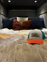 Inside a comfortable RV rental: A cozy bed with pillows, a fluffy blanket, and a Nirvana Upfitters cap. Ready for adventure.