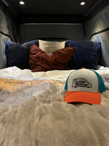 Inside a comfortable RV rental: A cozy bed with pillows, a fluffy blanket, and a Nirvana Upfitters cap. Ready for adventure.