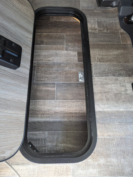 An open RV rental floor access hatch with a black frame, revealing wood-look tile flooring and a metal tie-down point.