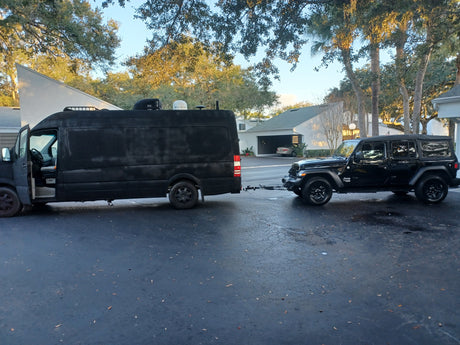 Black RV rental Sprinter camper van flat-tows a matching Jeep Wrangler on a sunny driveway, ready for adventure.