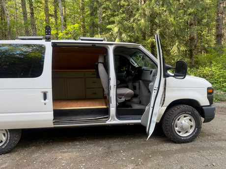 Open white camper van, showcasing custom wood interior & cabinets, parked in a lush forest. Perfect for an rv rental adventure.