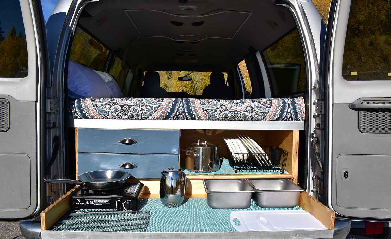 This rv rental features a cozy bed above a pull-out kitchen with stove, pan, and dishes, perfect for camper adventures.