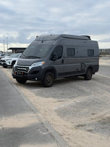 Off-road ready dark gray Ram ProMaster camper van, an ideal RV rental, parked on sand near a road under a cloudy sky.
