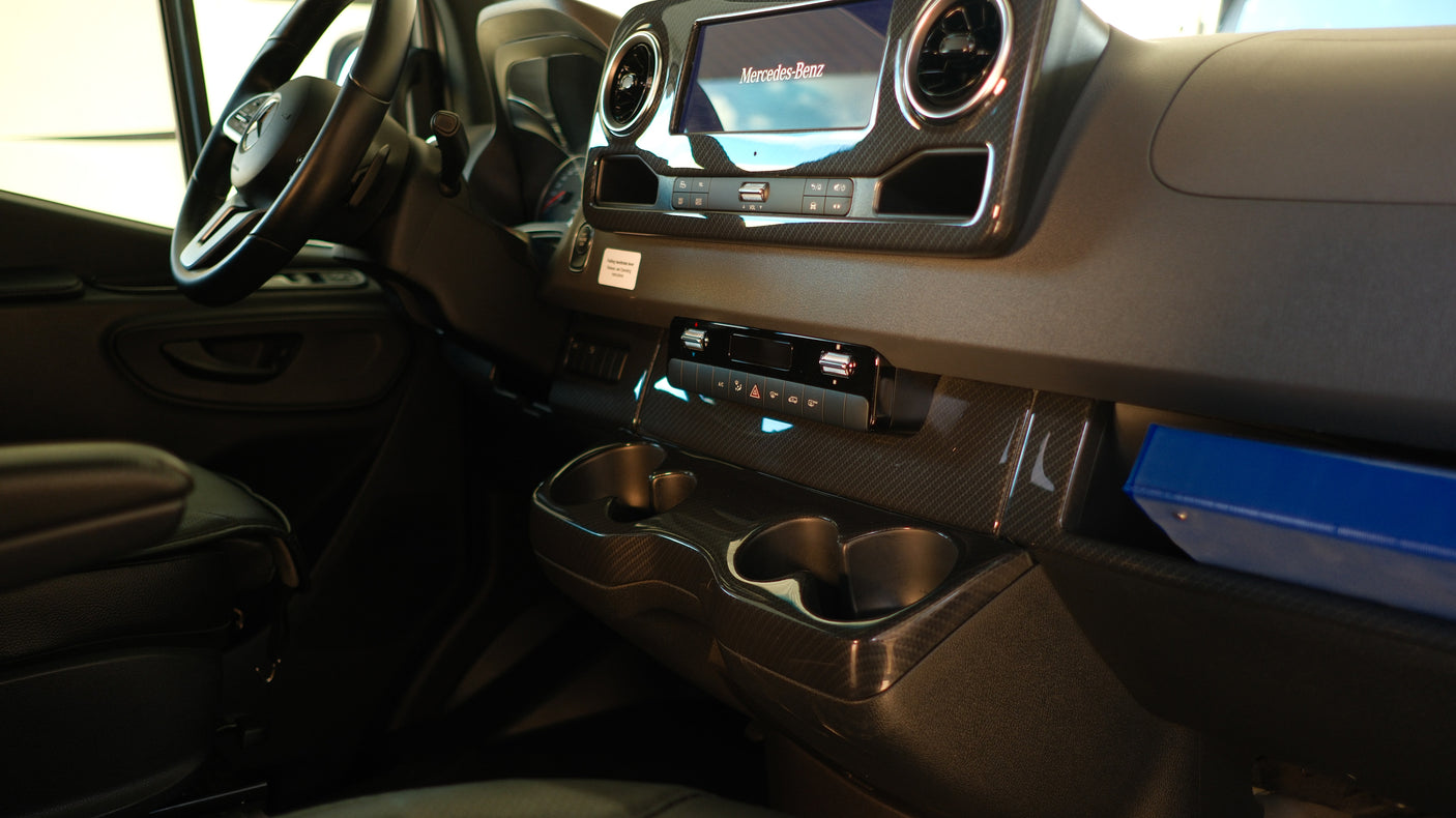 Inside a Mercedes-Benz RV rental: dashboard with steering wheel, infotainment, controls, cup holders, and carbon-look trim.