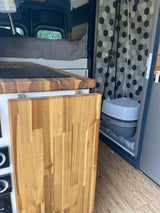 Compact RV rental interior featuring a bed, kitchen counter with cooktop, and a convenient portable toilet in the shower.