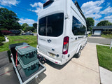 White Ford Transit RV rental parked on a suburban driveway, with gear secured on a rear cargo carrier under a sunny sky.