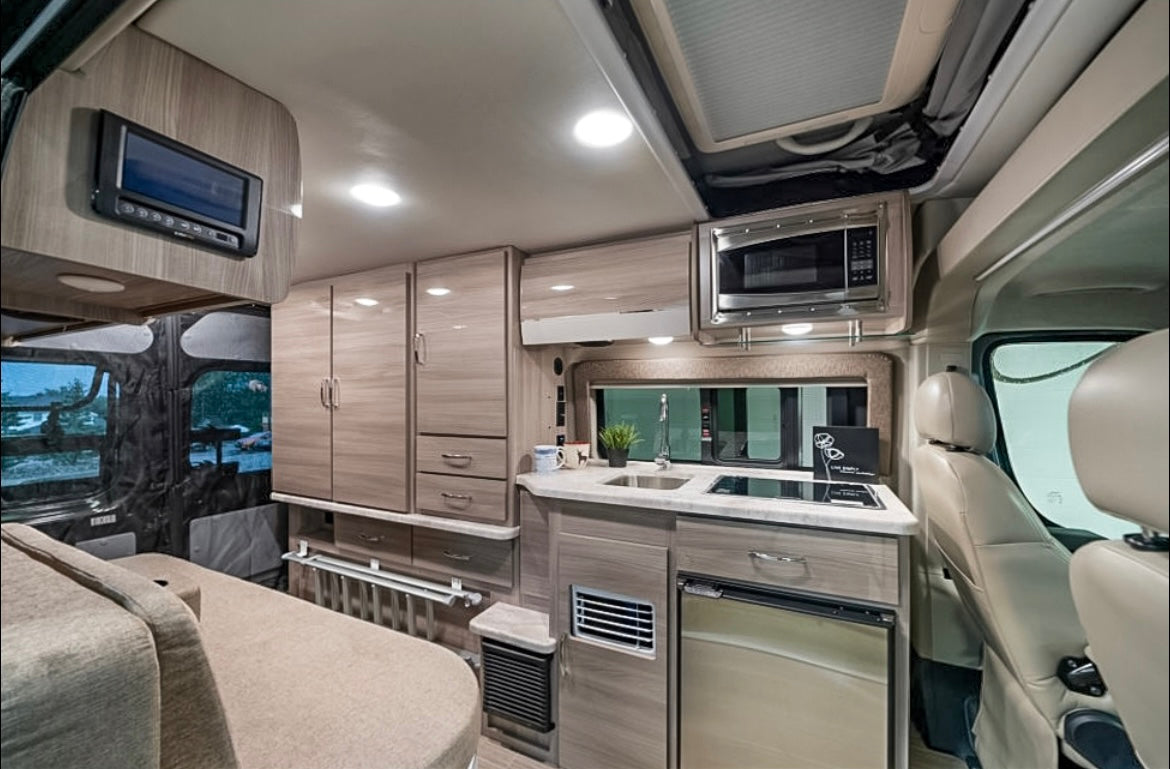This modern RV rental interior features a compact kitchen with a sink, microwave, fridge, and convertible bed/seating area.