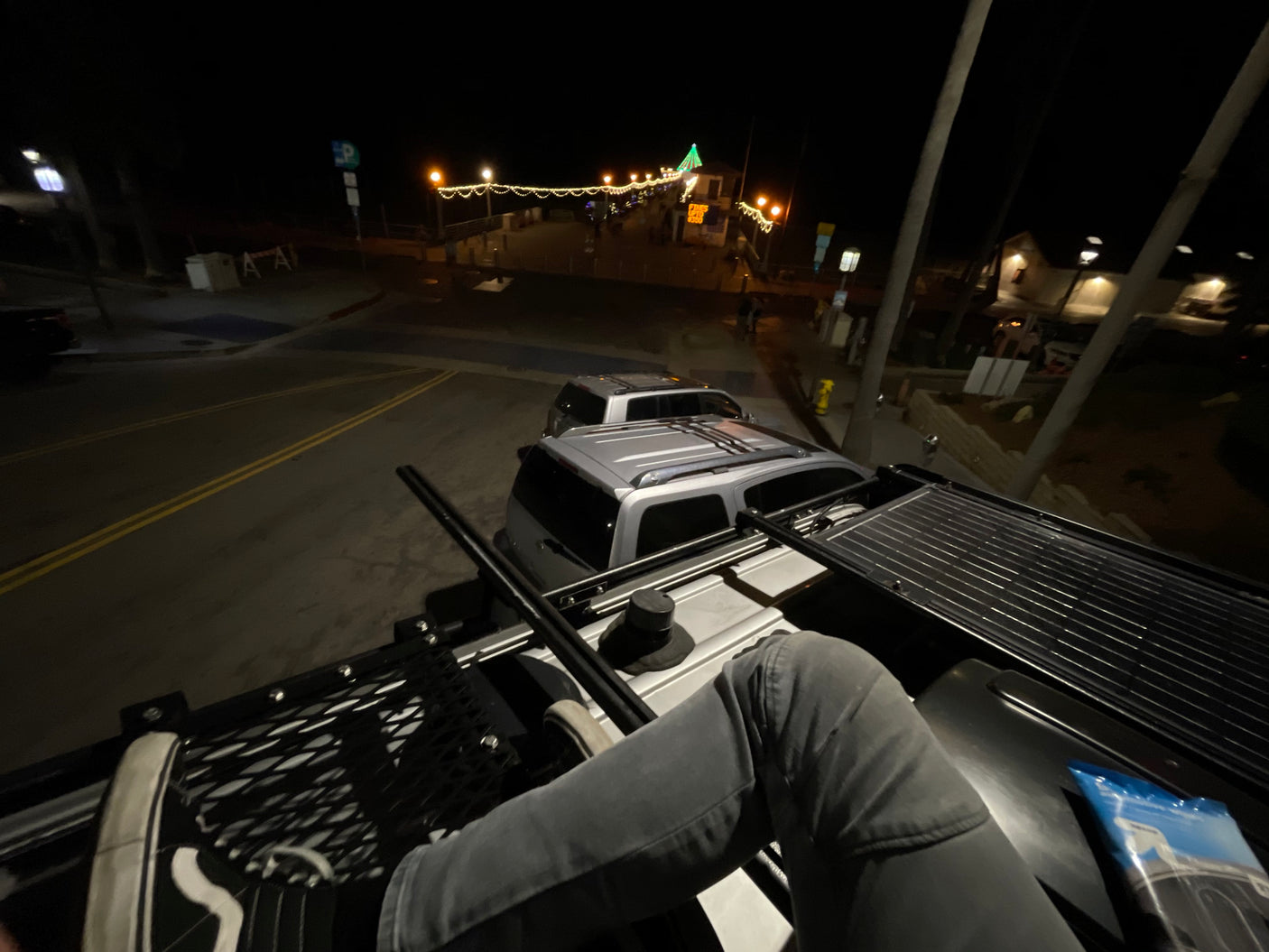 Night view from an RV rental rooftop, capturing a lit coastal pier, street, and parked cars below.