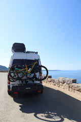 White rv rental van with mountain bikes on a rack, overlooking a vast blue lake from a scenic road. Ready for adventure!