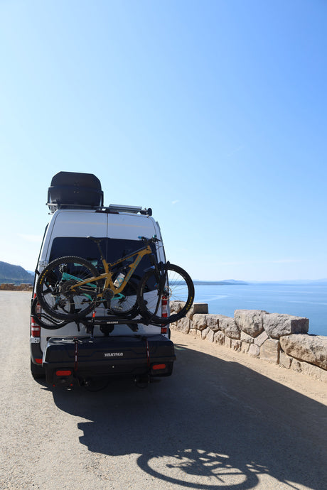 White rv rental van with mountain bikes on a rack, overlooking a vast blue lake from a scenic road. Ready for adventure!