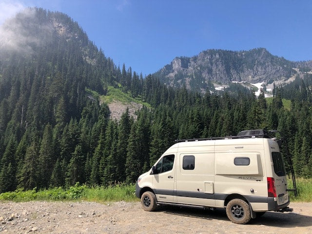  surrounded by a lush forest under a blue sky.|Cozy RV rental: Person relaxing in a brightly lit camper van bed at dusk with rear doors open.|A cream-colored RV rental van is parked on a dirt road amidst majestic mountains — photo 5