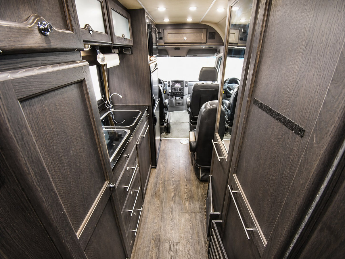 Explore this modern RV rental interior featuring a compact galley kitchen, dark wood cabinetry, and a view to the driver's cabin. — photo 11