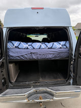 High-top van rv rental: converted interior with elevated bed & storage, seen through open rear doors. Tow hitch visible.