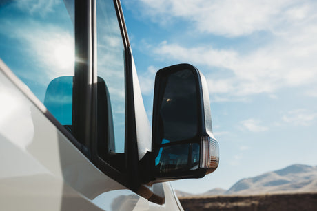 Your RV rental journey awaits! A white van's side mirror and window reflect a sunny sky. Distant mountains beyond.