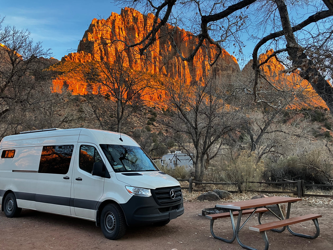 White Sprinter RV rental van set up at a campsite with a picnic table and majestic orange-lit mountains.