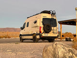 Rear view of a cream Winnebago Revel rv rental van with roof rack and ladder, parked in a sunny desert landscape.