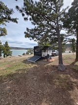 Enjoy nature with this RV rental! A travel trailer with a ramp, truck, chairs, and pine trees by a serene lake.