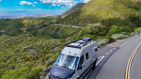 Gray RV rental parked on a scenic, winding mountain road, with lush green hills, blue sky, and a distant cyclist.