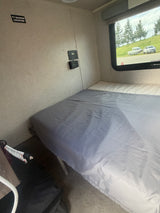 RV rental bedroom with a made bed, wall-mounted controls, and a window view showing a truck, boat, and trees.