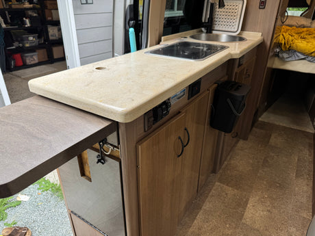 This rv rental interior features a compact kitchen with a sink, stovetop, extendable counter, and adjacent cozy bed.