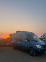 Scenic RV rental: Blue Sprinter van at sunset over the ocean, bikes on rear. Ideal for adventure.