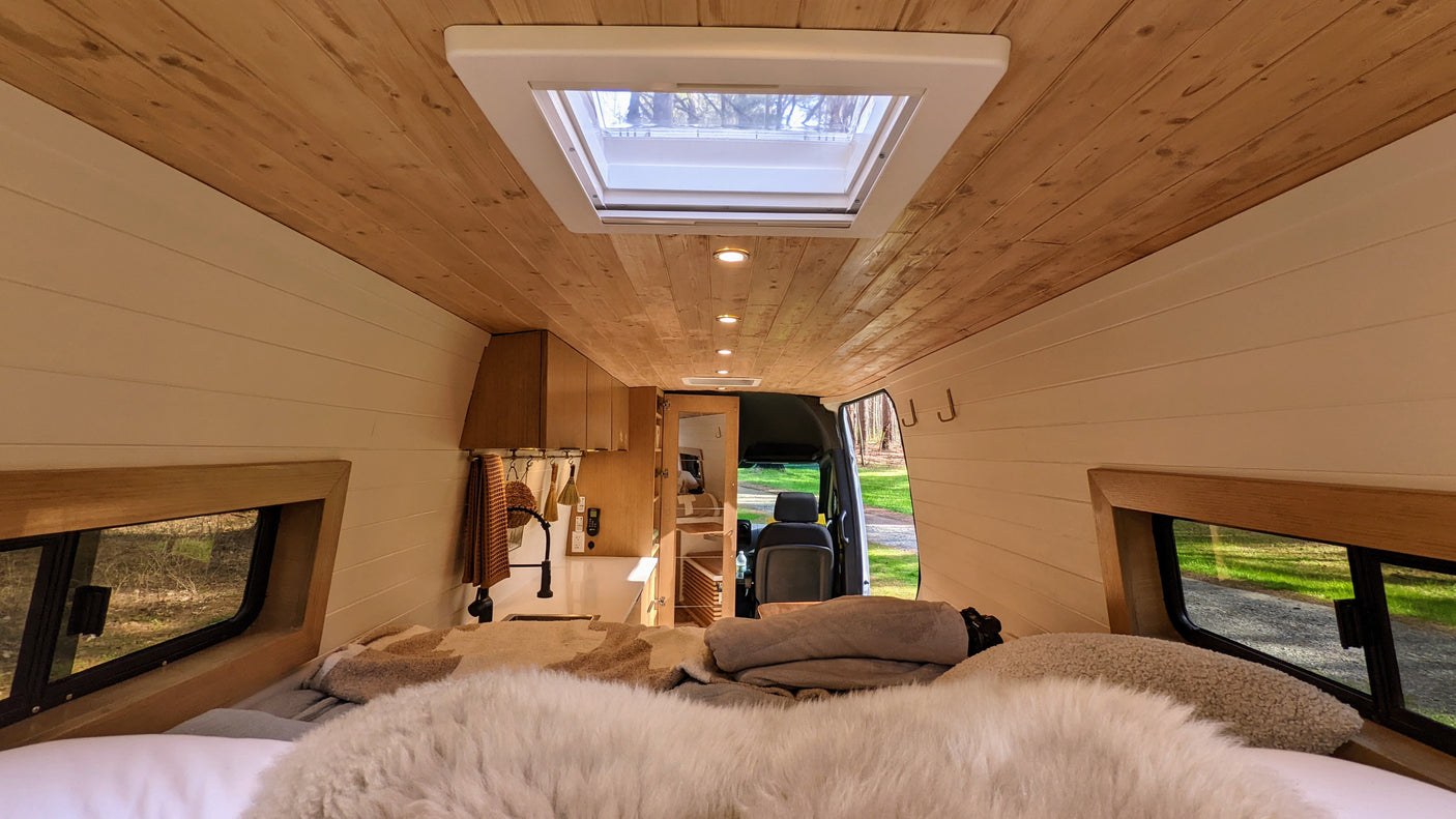 Cozy rv rental interior: view from the bed with fluffy blankets, showcasing a wood ceiling, skylight, and galley kitchen.