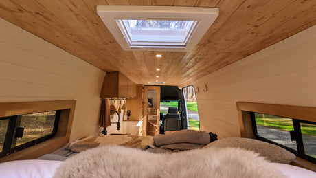 Cozy rv rental interior: view from the bed with fluffy blankets, showcasing a wood ceiling, skylight, and galley kitchen.