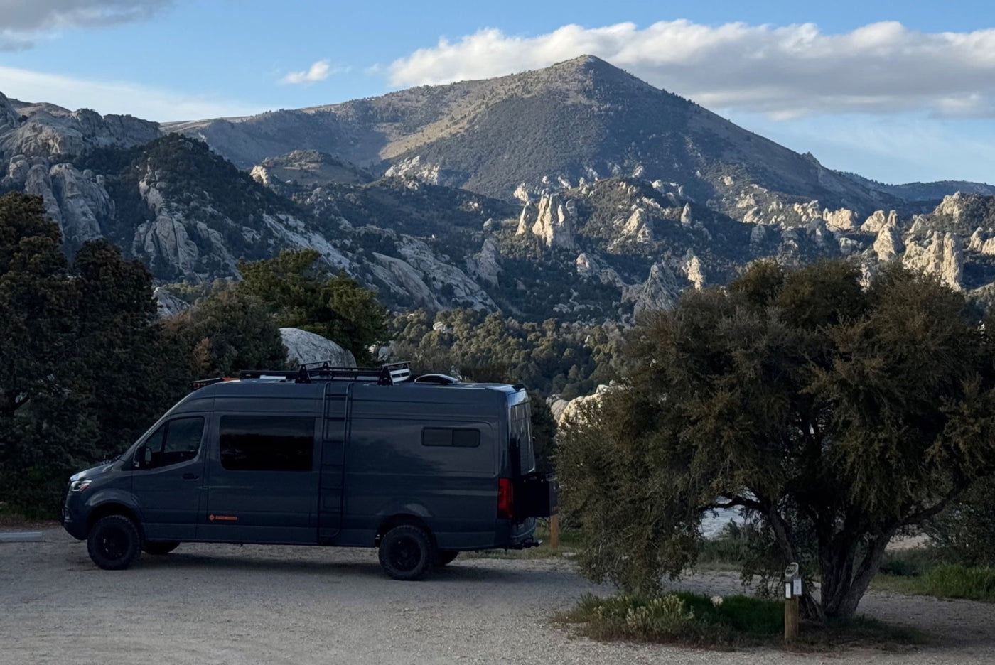  parked on gravel with mountains & trees in the background.|This dark camper van RV rental is ready for adventure! Parked in a stunning mountainous landscape with rocky peaks & trees.|Dark gray Mercedes Sprinter rv rental camper van with off-road tires and light bars