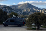 This dark camper van RV rental is ready for adventure! Parked in a stunning mountainous landscape with rocky peaks & trees.