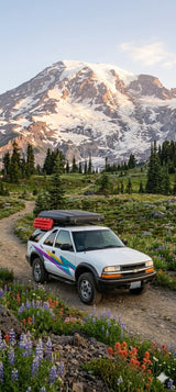 White adventure SUV with roof gear on a mountain path amidst wildflowers and a snowy peak. Ideal for an outdoor rv rental.
