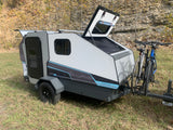 This sleek rv rental teardrop camper, complete with a bike rack holding two bicycles, sits ready for outdoor adventure. Its rear hatch is open.