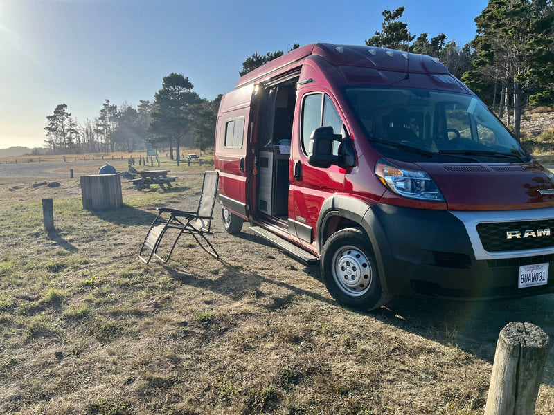  with a blue outdoor mat laid out and a mountain bike nearby.|Scenic rv rental views! Red Winnebago Solis camper van by a serene mountain lake with a rocky peak.|Cozy interior of an RV rental rooftop tent with a comfy bed — photo 18
