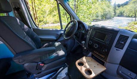 Spacious RV rental van interior with driver's seat, dashboard, and custom tray. Enjoy riverside camping views.