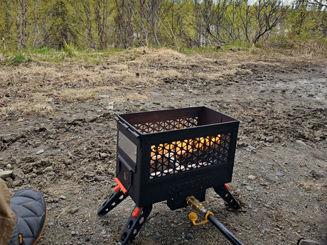 A black portable propane fire pit with orange legs burns brightly on a gravel campsite, perfect for an RV rental outdoor adventure.