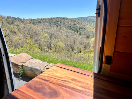An RV rental provides this stunning spring mountain view. Light green trees fill the valley, framed by a cozy wooden interior.