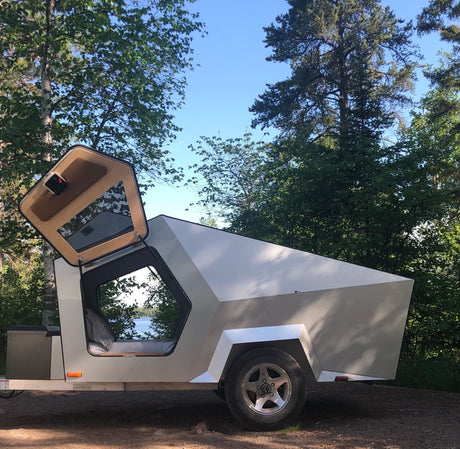 Unique, angular compact RV rental by a lake. This modern travel trailer has an open door, revealing a cozy bed inside.