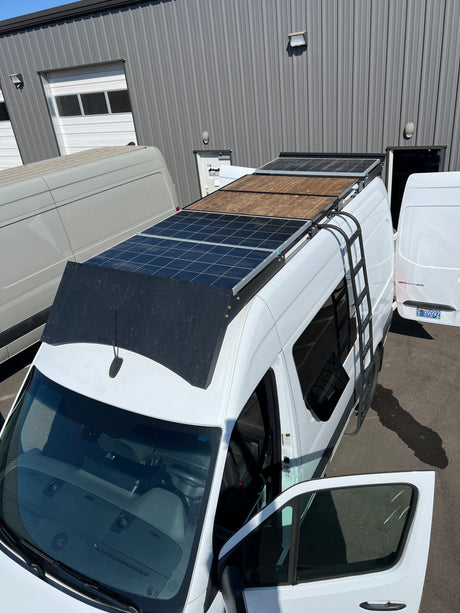 Overhead view of white Sprinter camper van with solar panels & ladder, ideal RV rental, parked by a building.