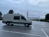A grey RV rental van with roof gear, parked on a wet day, overlooks the foggy Golden Gate Bridge.