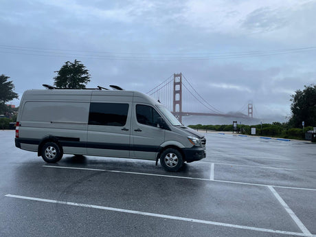 A grey RV rental van with roof gear, parked on a wet day, overlooks the foggy Golden Gate Bridge.