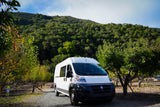 White RV rental van parked on a gravel path in a sunny orchard, with a lush green mountain backdrop. Scenic nature escape.