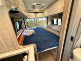Inside an RV rental: a cozy bedroom with a blue blanket, folded towels, and rear windows, plus a compact kitchen setup.