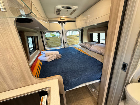 Inside an RV rental: a cozy bedroom with a blue blanket, folded towels, and rear windows, plus a compact kitchen setup.